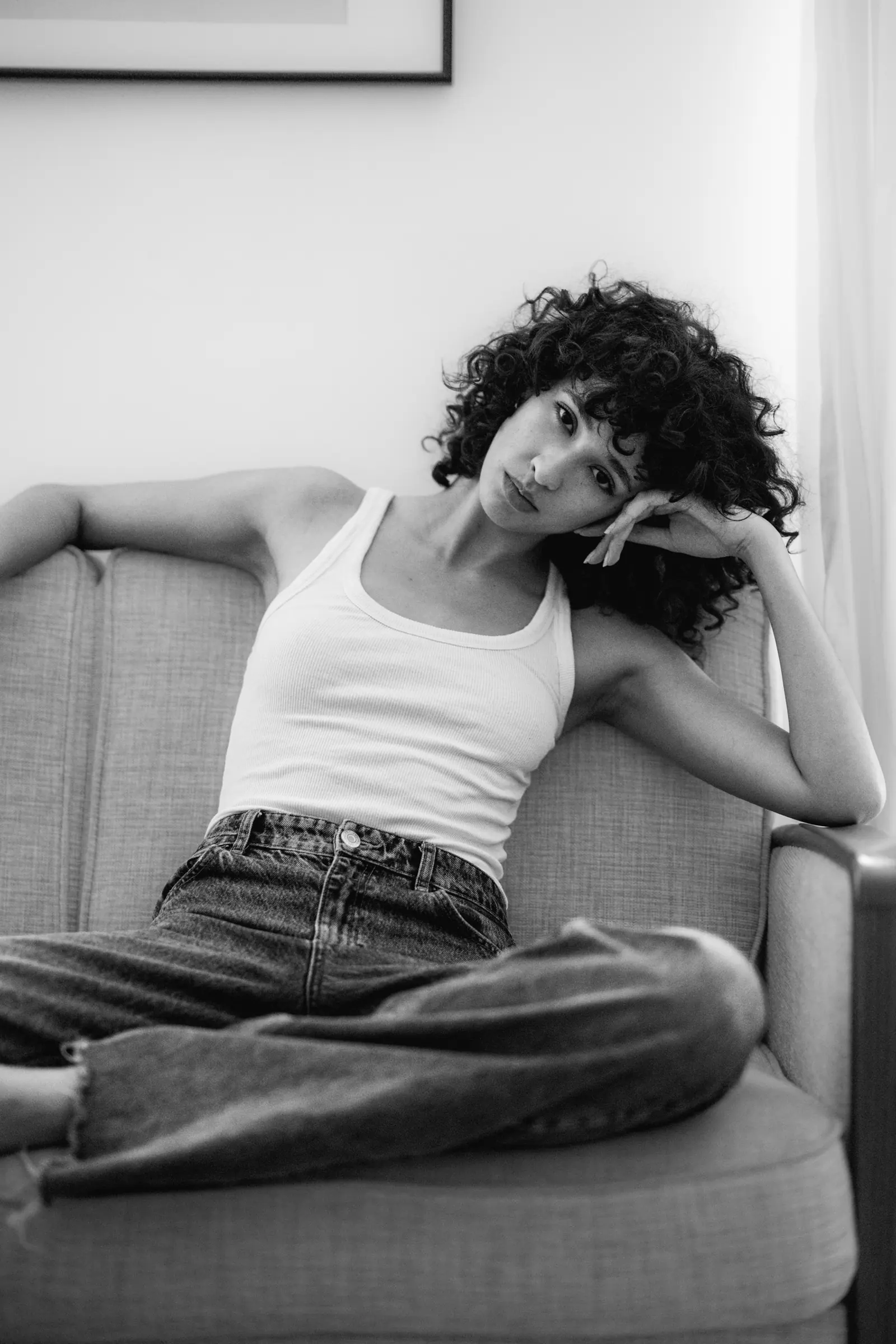 Woman with curly hair in a white vest, leaning on a sofa, in black and white