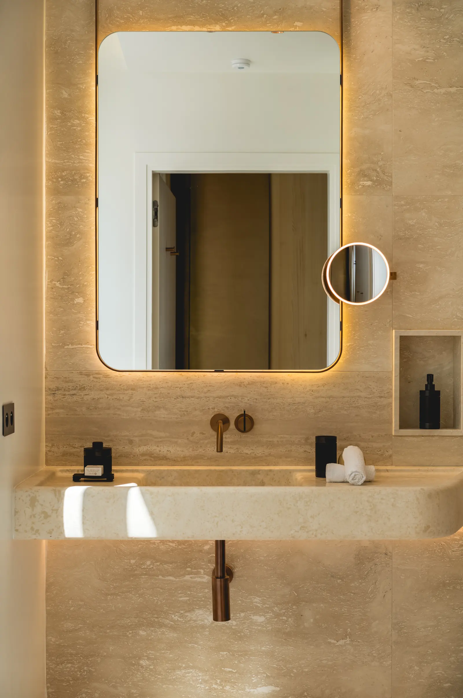 Backlit mirror above a travertine basin with bronze taps
